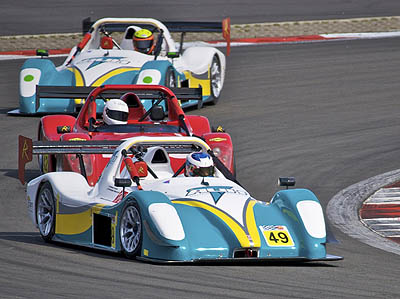 Sad News Pistonheads - The image is a dynamic photograph capturing a racing car competition on a track. The scene features two sports cars traveling swiftly around a negotiated curve. The lead vehicle is the most prominent, with a vibrant combination of white, blue, and yellow bodywork, while the trailing car has white and red hues. Both vehicles display the number "49" in prominent yellow text, indicating a specific entry in a race or event. The photo captures the high speed and aggressive behavior of these cars as part of competitive motorsport.