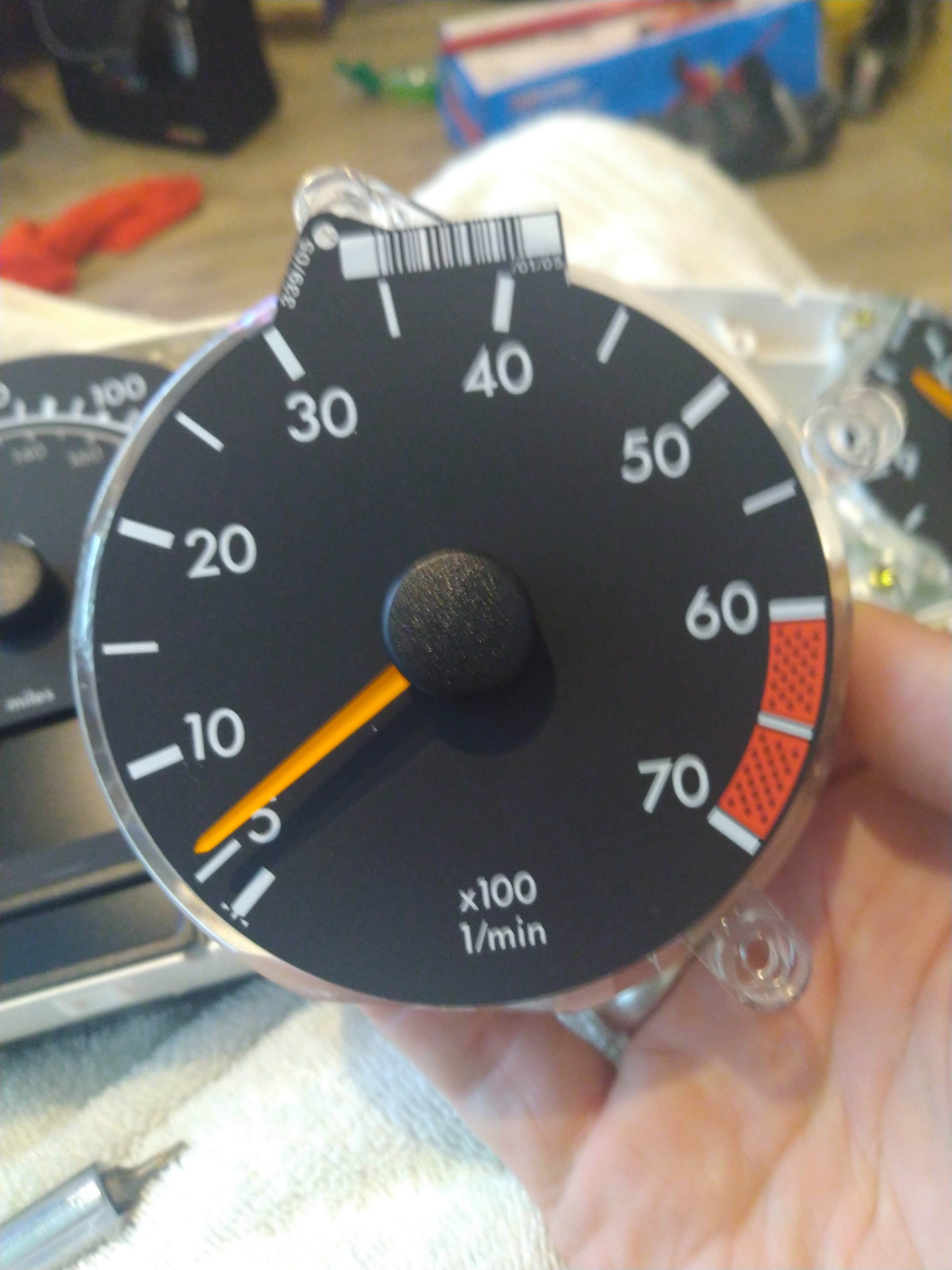 A close up of a person holding a pair of scissors - Pistonheads - The image shows a person holding a gauge that reads "0.1", indicating a low battery reading. This gauge appears to be part of a larger setup, possibly related to electronics or automotive components given the presence of what seems to be a small car model and a speedometer needle. There is a human hand visible at the bottom of the frame, holding the gauge in place. The background suggests an indoor setting with some household items scattered around.