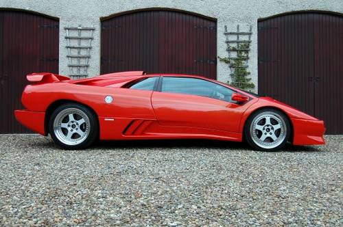 Pistonheads - The image shows a sleek red sports car parked in front of brown garage doors. The car is positioned diagonally, highlighting its angular design. It features a prominent front spoiler, side air intakes, and a large rear wing, which are all characteristic of high-performance vehicles. The setting appears to be an outdoor area, as indicated by the natural light and the ground covering. The color of the car contrasts sharply with the brown hue of the garage doors, drawing attention to the vehicle.