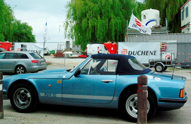 Three TVR S Series succeed at Battle of Waterloo - Page 1 - S Series - PistonHeads - The image focuses on a classic car that is predominantly blue with a white stripe. The car is parked against a fence that has a red barricade. In the background, there are several other vehicles, including trucks and cars, and a building with prominent signs for DuJoing and Dupiag. The sky is clear, indicating good weather. The environment gives off a vibe of a roadside business area with a picturesque setting.