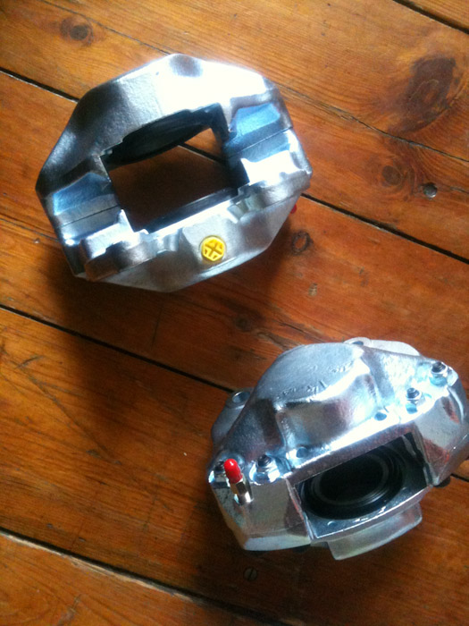 Refurbishment Brake Ate Pistonheads Caliper Recommendations - The image features two metallic objects lying on a wooden floor, possibly car parts given their metallic texture and automotive appearance. The parts appear to be dark in color, with one showing a yellow and black sticker and a red-painted part with a bolt and a thumb screw. The floor has visible wooden planks with noticeable creases and gaps in between, creating a rustic background. The lighting suggests an indoor setting with artificial light illuminating the objects. There are no texts or additional objects in the frame. The photograph's composition is centered with the metallic parts occupying most of the frame.