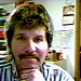 The image shows a man who appears to be indoors, possibly in a room with a wall clock visible in the background. The man is holding his chin with his hand, which is resting on his chest. He has a thick, curved mustache and his hair is a mix of brown and gray. He seems to be wearing a patterned shirt. There's a soft blur around him that suggests the background might be out of focus.