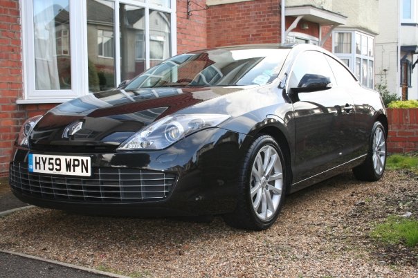 Coupe Laguna Pistonheads - The image depicts a black car parked on a gravel driveway in front of a brick house. The car is a coupe-style vehicle, indicated by its sleek lines and design. It has a distinctive emblem on the grille, suggesting it is a specific model. There are windows visible in the background, possibly indicating that the car is parked in the vicinity of the house, which could be a point of interest for the car's owner or a neighborhood resident. The overall setting suggests a residential area with houses that have ornamental features like brickwork and possibly white frames for windows.