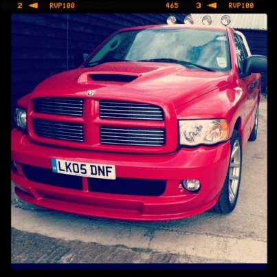 Just bought a towing vehicle! - Page 1 - Vipers - PistonHeads - In the image, we see a vibrant red Dodge Ram pickup truck parked on a concrete surface. The truck, with its black grill and gold hood, gleams under the light. The image is taken from a slightly angled perspective, giving a full profile view of the truck. A license plate reading "LK05 DNF" adorns the front bumper. A set of four off-road lights are mounted on a rack atop the truck, adding to its rugged appeal.