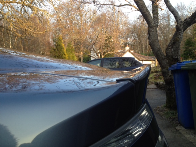 DBS wheels on DB9 - (more or less) - Page 1 - Aston Martin - PistonHeads - The image depicts a residential scene with two parked cars. The foreground features a dark blue car, likely a sedan or a wagon, characterized by noticeable reflections on its hood, suggesting a glossy and well-maintained surface. The spill of light on the car's hood highlights its condition and might indicate that this is a recently cleaned or waxed vehicle.

In the background, partially obscured by the larger car in the foreground, is a white house with a uniform appearance that aligns with a typical suburban home. To the right of the frame, there is a blue trash can, reinforcing the domestic setting.

The setting appears to be during daylight, with natural light illuminating the scene. Trees without leaves suggest that the photograph was likely taken in late autumn or winter. There's no immediate evidence of snow on the ground, but the lack of foliage implies cold weather.