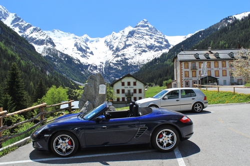 First  Boxster - Page 1 - Boxster/Cayman - PistonHeads - The image displays a picturesque landscape with two parked cars resting on a slope with a distant view of a mountain range. The foreground shows a black, two-doors car with its top down, parked under a clear blue sky, while a slightly smaller silver car is positioned sideways behind it. Behind the cars, a quaint white building with a combination of timber and stone construction is nestled at the base of a steep incline. The buildings and cars are surrounded by lush greenery, contributing to the serene and beautiful setting.