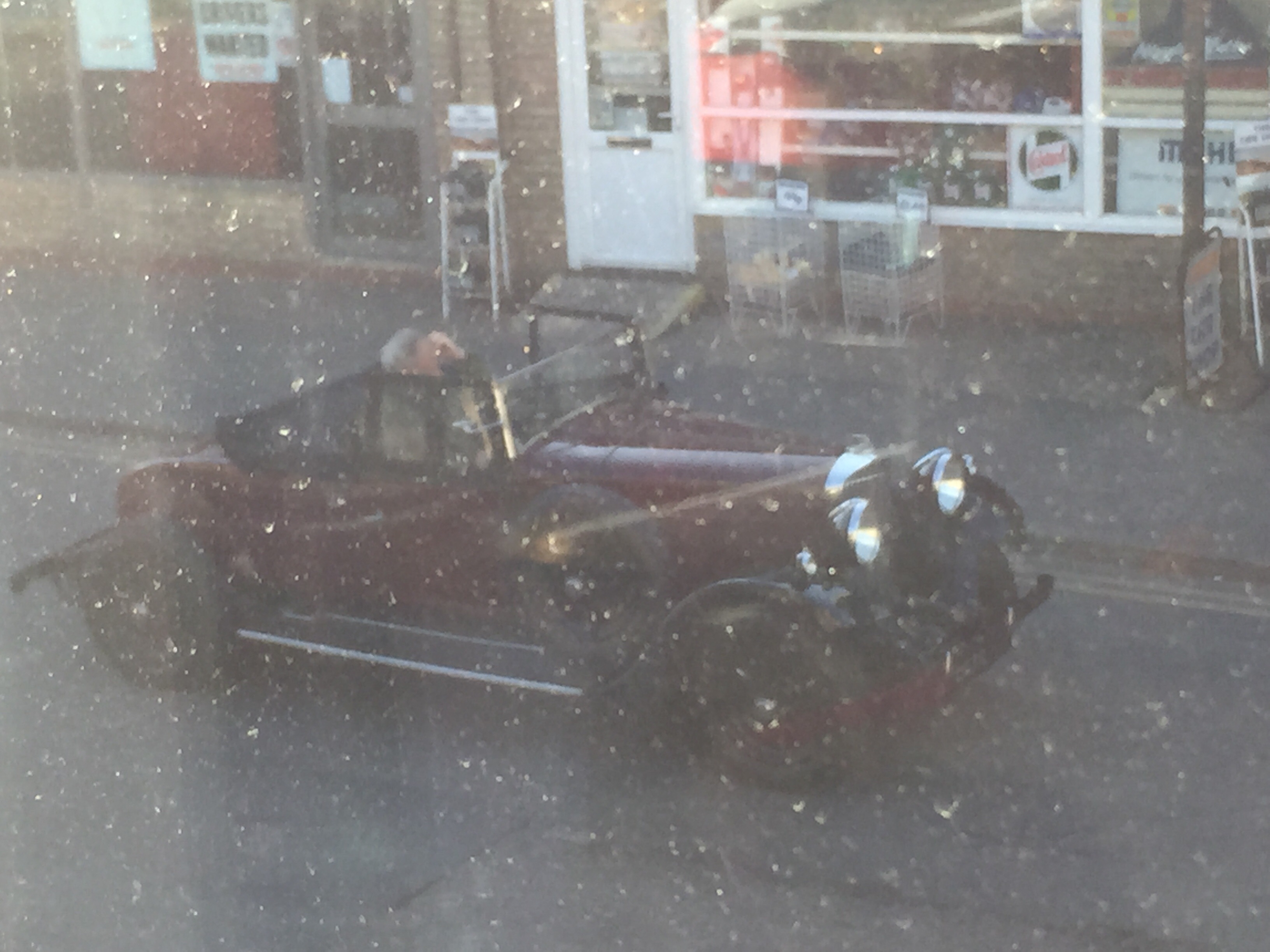 Herts, Beds, Bucks & Cambs Spotted - Page 366 - Herts, Beds, Bucks & Cambs - PistonHeads - The image captures an open old-time car parked on the side of a road, which leads up to a small store. The car, likely a classic or vintage model, has a glossy black finish and features a bucket seat design. A person is partially visible through the driver's side window, but their face is obscured by the car's reflection on the glass. The store in the background displays merchandise in its window and is shrouded by the car's shadow. The scene exudes a sense of nostalgia and leisurely pace, transporting viewers back to the era of classic automobiles.