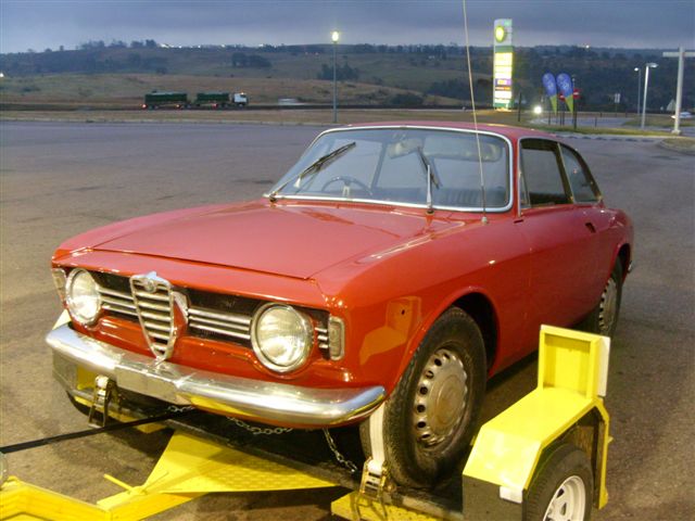 Beautiful Pistonheads Bertones - The image shows a red vintage car, prominently featuring a luggage carrier or luggage rack attached to its rear. The car is parked on a concrete surface, positioned in a location that suggests it might be near an airport or a transit hub. The setting sun casts a warm glow on the vehicle and equipment. The scene conveys a sense of travel or a journey, particularly for those familiar with air travel and its pre- and post-flight vehicles.