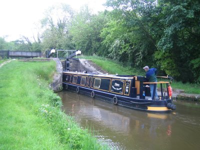 The image showcases a tranquil river scene, featuring a small two-deck canal boat in motion. The boat, with its brown and black color scheme, is being navigated by a single person - the skipper - who is standing on the upper deck of the boat. The boat is moving along the canal, which is flanked by a lush green bank and an overhanging canopy of trees, providing a serene and natural backdrop to the scene. The surrounding landscape is vibrant and verdant, creating a peaceful and idyllic atmosphere.