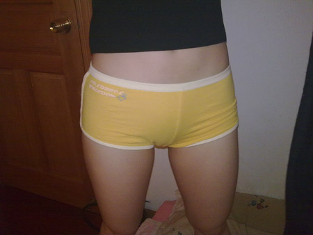 The image shows the lower body of a person wearing yellow-colored underwear with a white waistband. The underwear brands appear to be "PRIESTLY HAPPIN" and "Undies Re" or "Re Re Undies". The person is standing indoors, and a portion of a wooden door can be seen to the left of the person. It appears to be taken in a bedroom with a light-colored wall visible behind the person. The photo has a casual and domestic feel.