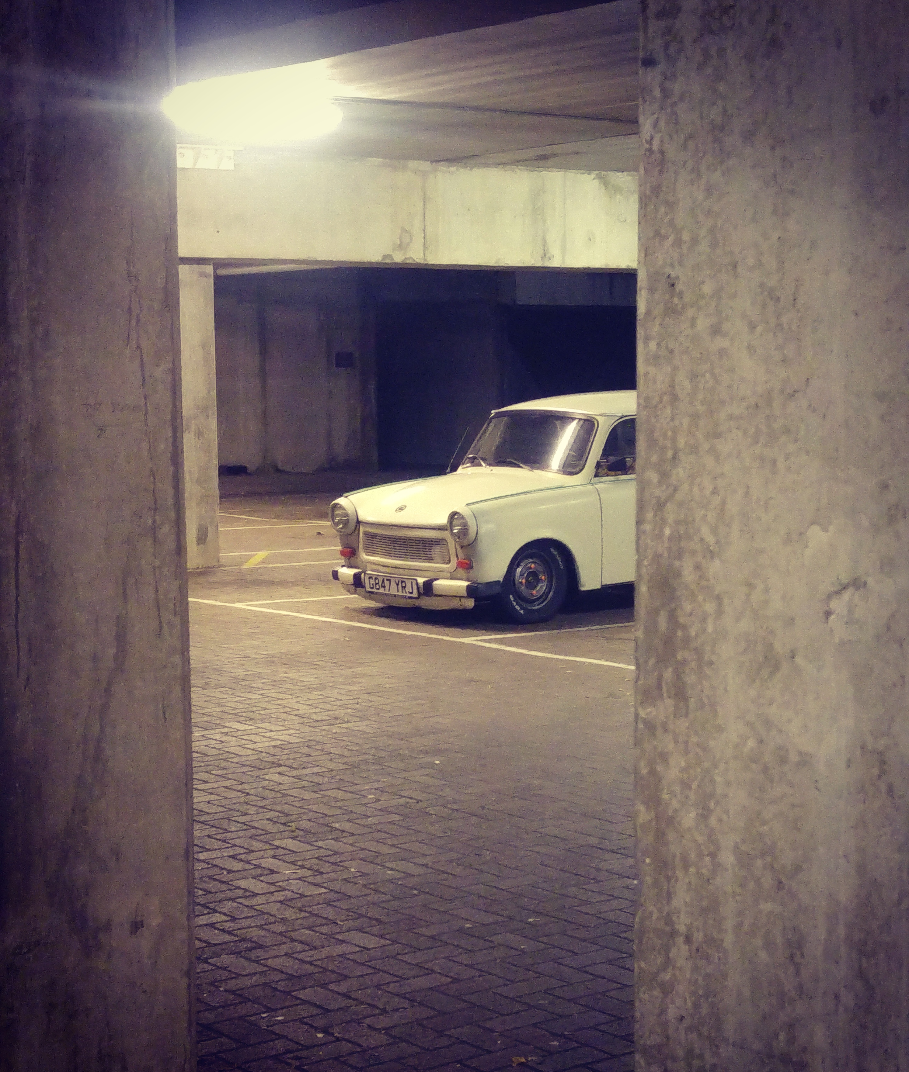 Lets see a picture of your classic(s) - Page 368 - Classic Cars and Yesterday's Heroes - PistonHeads - The image features an old, white car parked in a shaded underground parking lot. The car's vintage design is contrasted by the modern-looking brick floor beneath it. The sunlight coming in through gaps in the ceiling illuminates the car, highlighting its worn-out features. The perspective of the photograph is from between two stone pillars, looking out towards the car.