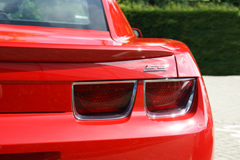 Pistonheads Import Camaro - The image displays the rear view of a bright red sports car. The car is parked, with the pavement visible under it. The tail lights are clearly visible, showcasing their sleek design with a horizontal fin on the left side. The car's shiny surface reflects light and its design suggests speed and performance. The image captures the car outdoors, possibly during the warmer months, as indicated by the surrounding greenery.