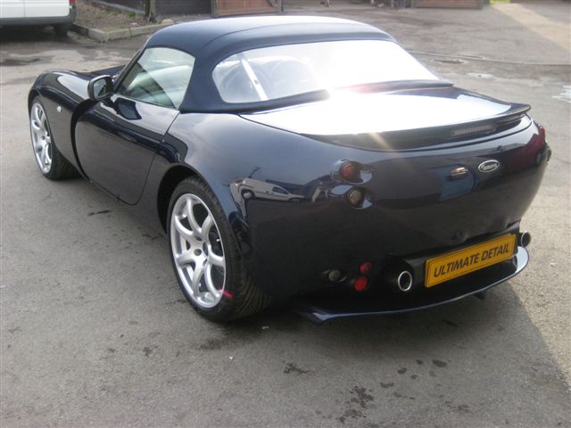 Cambridge Ultimate Pistonheads Detail - The image shows a sleek black sports car parked on a concrete surface. The car's top is down, revealing an interior presumably lined with accent foam in a contrasting blue and red pattern. The vehicle features two doors and has a spoiler on the rear, sporting a yellow "ultimate detail" license plate. Surrounding vehicles and some blurred figures in the background suggest this photo may have been taken in a public space like a parking lot or a driveway.