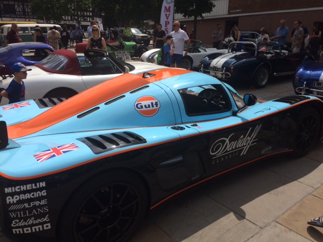 Ultima GTR - Gulf Livery - Page 1 - Ultima - PistonHeads - This image captures the allure of a car show. The gathered crowd, some close to the camera, some further away, are all attentively observing the stunning vehicles on display. The most eye-catching vehicle is a blue and orange GULF-DavCodec racing car, its American flag decoration proudly displayed. The scene is set in a parking lot, which appears to be filled with many more vintage and racing cars, adding to the overall ambiance of the car show.