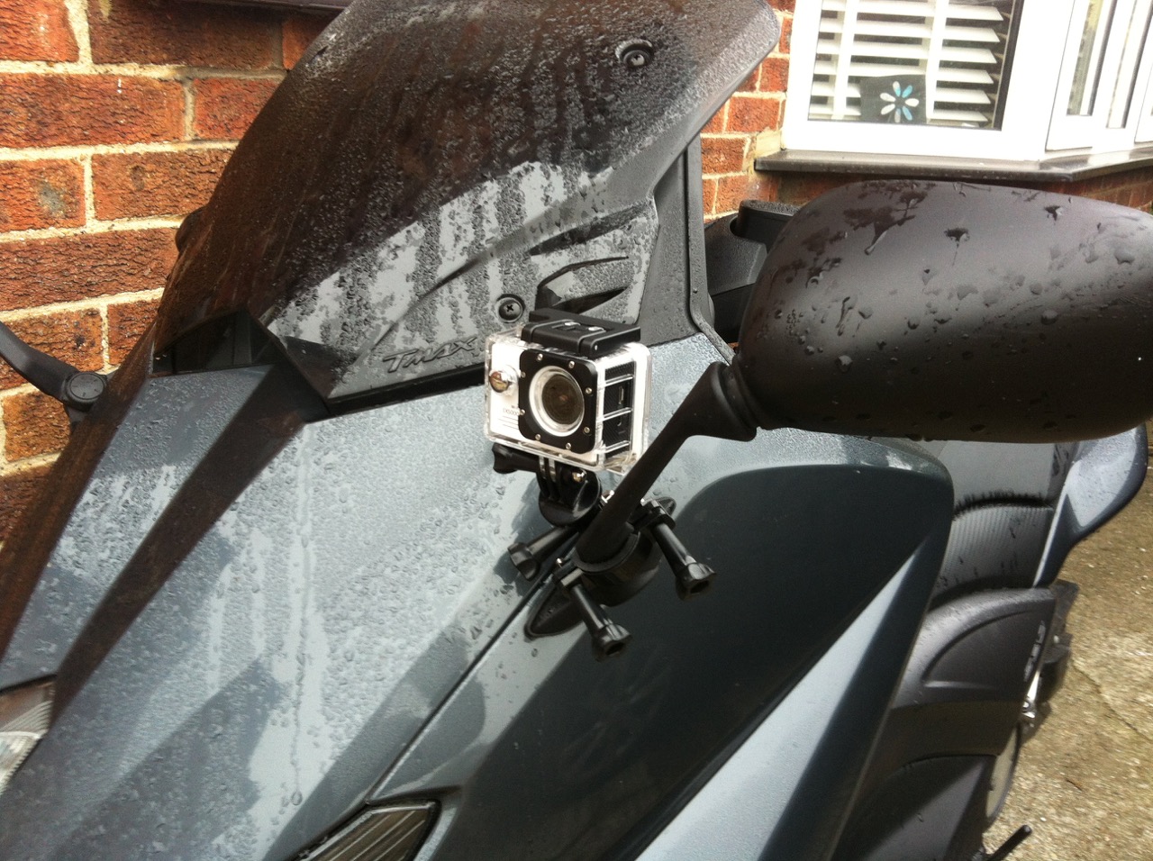 Bargain Crazy Thread - Page 60 - Biker Banter - PistonHeads - The image displays a black motorcycle parked against a brick wall. Attached to the handlebar is a camera, secured with a bracket. The surface of the motorcycle appears wet, suggesting that it may have been ridden in the rain. The motorcycle is made of metal, and there is a reflector on the back, indicating that the rider likely values safety. The design of the motorcycle is contemporary, with curves and lines that give it a sleek appearance.