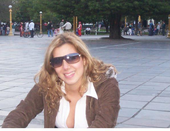 Efectodrago - The image is a photograph featuring a woman who appears to be smiling and posing for the camera. She has shoulder-length curly hair, is wearing sunglasses and a brown jacket with a fur-trimmed hood. Her face is partially obscured by the sunglasses, and the woman is looking directly at the camera.

She is seated outdoors on a paved surface that could be part of a plaza or a sidewalk. The background of the photo shows a confined green space that seems to be a city park, with several trees and pieces of street furniture.

People can be seen in the distance, though they are out of focus and do not appear to be directly interacting with the woman in the foreground. The lighting in the image is diffused, suggesting either overcast conditions or an area with plenty of shade. The overall atmosphere is casual and relaxed.