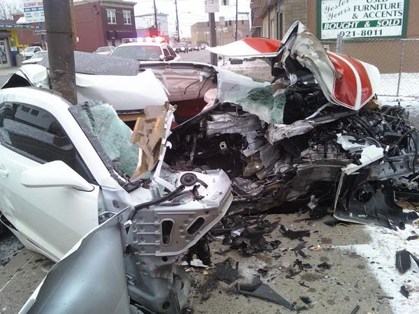 Pistonheads Camaro Waste - The image depicts a severely damaged car in what appears to be an urban environment. The car's front end is smashed in, suggesting it has been in a sizable collision. Visible behind the car, there is a horror store sign, indicating the presence of various commercial businesses. The car is mangled and in a state of disrepair, with various components hanging off.