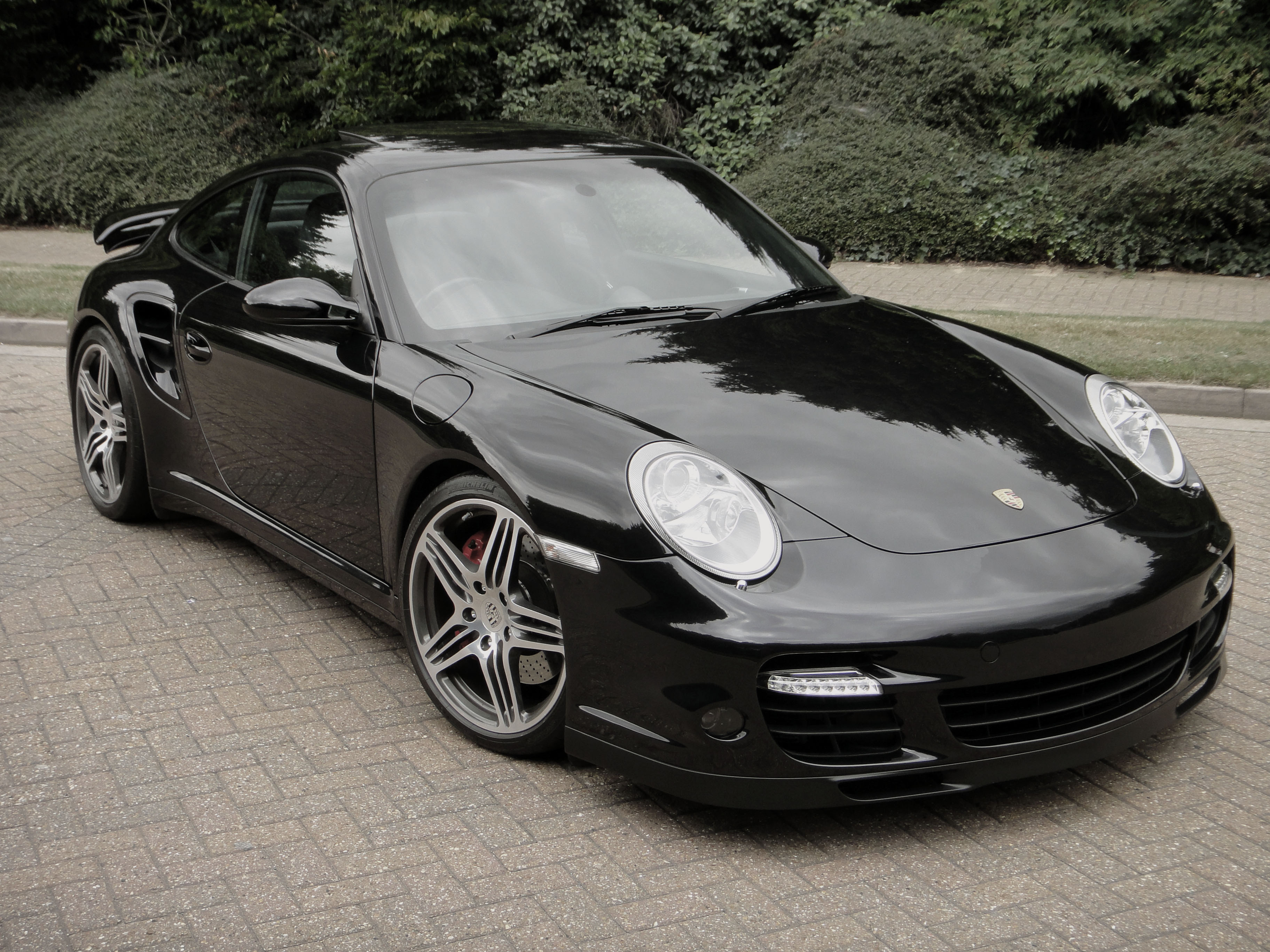 Pictures of 997 turbo's - Page 9 - Porsche General - PistonHeads - The image showcases a sleek black Porsche sports car, parked on a brick pavement. The car is positioned at an angle, with its front slightly off-center, revealing a glimpse of the passenger side. Its design is characterized by a large grill and prominent headlights, indicative of its sleek and sporty aesthetics. The vehicle's glossy finish and metallic accents add to its luxurious and high-performance appearance. The brick pavement on which it sits provides a textured contrast to the smooth surface of the car.