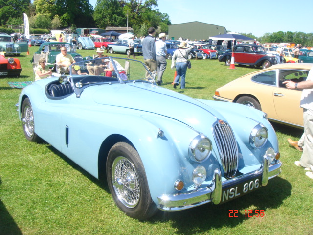 Old Warden car show - Page 1 - Classic Cars and Yesterday's Heroes - PistonHeads - The image showcases a classic blue Jaguar car positioned on a grass field, possibly a car show or exhibition. The vehicle, with its shiny metal body and white wheel rims, stands out prominently in the middle of the frame. In the background, the vibrant greenery contrasts with the blue of the car, and there are several people strolling around the area, indicating a public event. The photo captures a moment of leisure and conversation among the visitors in the wondrous outdoor setting.