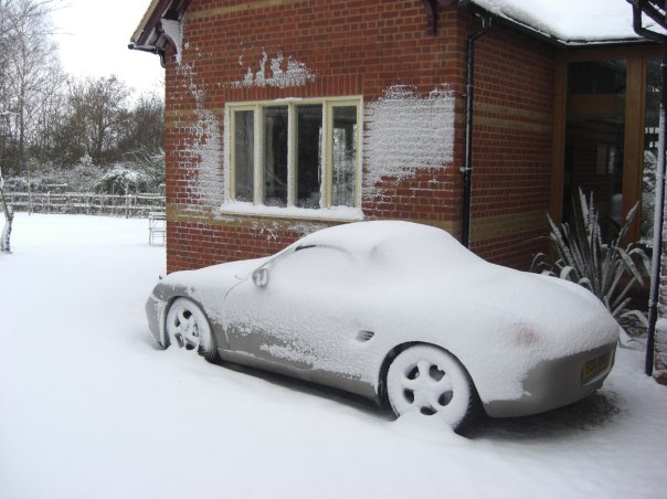 Pistonheads - The image portrays a residential scene with a car covered in snow. The car is a convertible, parked outside a brick building which features a black pipe running up the side. A black railing can be seen in front of the building, adjacent to a patch of snow-covered grass. A few bare trees are visible in the background, suggesting that the photo was taken during winter.