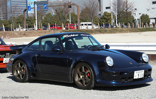 Better than a Speedline ? - Page 1 - Porsche Classics - PistonHeads - The image displays a dark-colored convertible car parked on a gravel area. The car is black with the top down, allowing an unobstructed view of the interior and the driver's side. The car features distinctive white stripes and a prominent rear fender with a slightly elevated black accent. The gravel surface beneath the car is uneven, and the photo is taken during the daytime under clear, bright conditions. In the background, there are other vehicles and structures, which suggest this could be a parking area or a driveway in a residential or commercial setting.