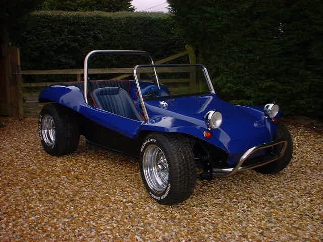 Owns Pistonheads Buggy Beach - The image showcases a vibrant blue single-seater European sports car parked on a gravel surface. The car has a sleek, low profile, with black walls and a transversal engine ribbon running along the side. Its wheels, incorporating grey hubcaps and continuing the car's black and silver theme, lend it a dynamic look. The background reveals a lush green hedge, creating a pleasing contrast with the car's bold color.