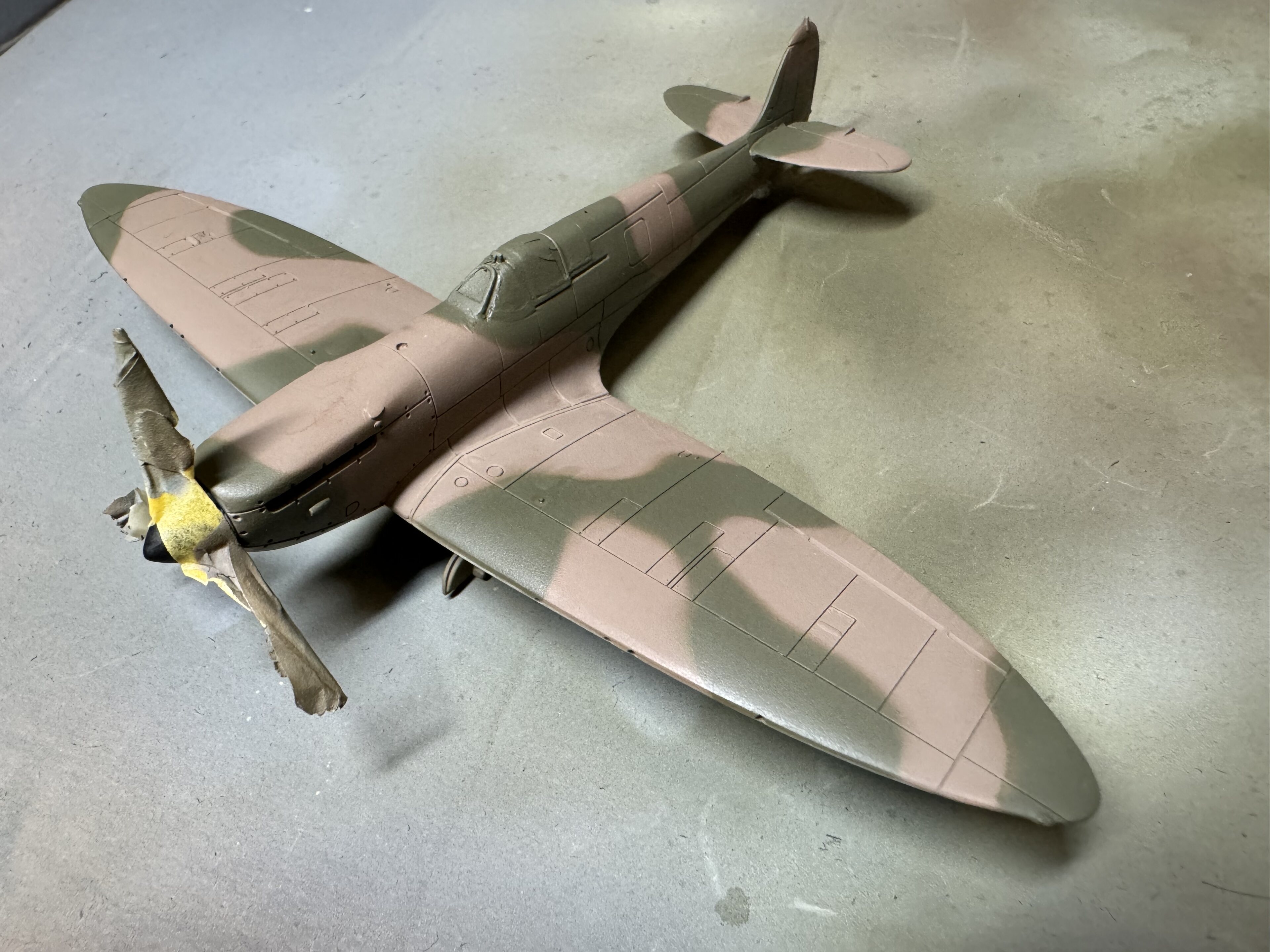 Pistonheads - The image shows a small model airplane, possibly a warplane, sitting on a surface. It is painted to resemble the camouflage patterns of a military aircraft, with shades of green, brown, and grey. The plane has a detailed design, with visible wings, a propeller, and other distinguishing features. In front of the airplane, there appears to be a small figure or model person, also dressed in camouflage clothing, giving the impression that they are piloting the aircraft.