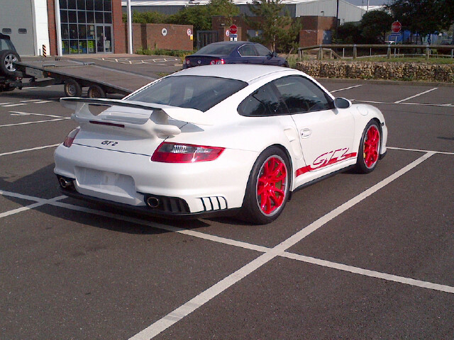 BEST 997 GT2 IN UK - How Much??? - Page 1 - 911/Carrera GT - PistonHeads - The image shows a white Porsche with a prominent rear wing, parked in a parking lot. The Porsche's design includes red detailing and wheels, and it's monogrammed 'GT1' on its side. The area around the car features brick buildings, suggesting an urban setting. In the background, other parked cars are visible, and a sign with a round red and white top can be seen.