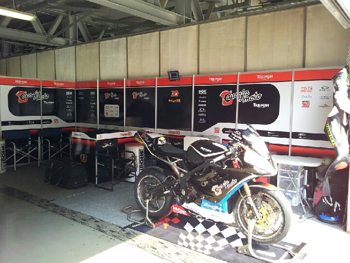 A row of motorcycles parked next to each other - Pistonheads - This image captures a brightly lit indoor space, likely a garage or workshop, with a sleek motorcycle placed on a checkered paddock mat, a common type found in racing pits. The motorcycle is black with blue and yellow details, and it's parked next to a pristine red and white motorsport wall with logos, implying a racing theme. Silver shelves stocked with motorsports equipment are also visible in the background, suggesting this space is well-equipped for motorcycle maintenance and racing preparation. The lighting is artificial and appears to be overhead fluorescent lamps, casting shadows and highlighting the various elements in the room.