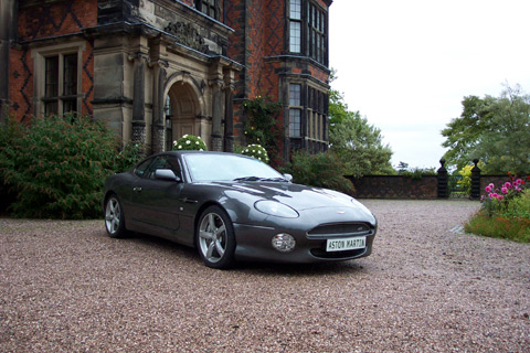 Your DB7 pictures. - Page 1 - Aston Martin - PistonHeads - The image features a parked sports car. The car is sleek and black, with a sunroof. It is parked in front of a majestic building that appears to be a mansion or large estate. The mansion has an ornate facade and is made of red brick. The car and the building are surrounded by a beautifully landscaped garden with flower beds and well-maintained shrubbery. The sky is clear and blue, suggesting that the photo may have been taken on a bright, possibly sunny, day.