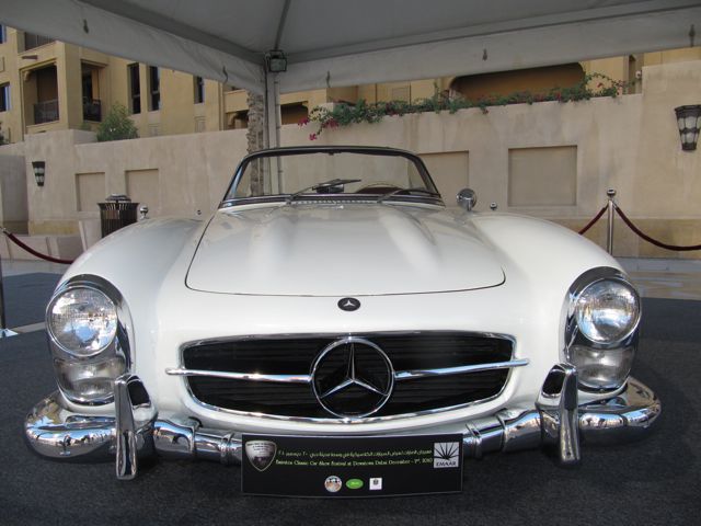 Emaar Pistonheads Classic - The image shows a classic white Mercedes-Benz car displayed under a tent. The car has a sleek, elongated design characteristic of vintage models. It features a prominent Mercedes logo at the grille and is parked on a stand or platform, which is standard for displaying cars at auto shows. There is text underneath the car, likely providing information about the vehicle or the conditions for accessing or examining it. The setting appears to be an auto show or exhibition, given the presentation and the backdrop suggesting other cars or booths.