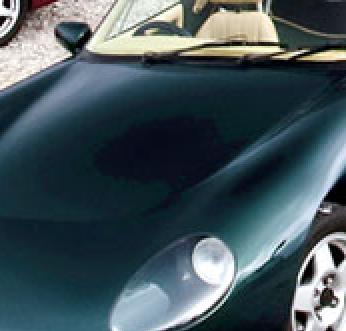 Tvrtastic Pistonheads Week - The image shows a close-up of the front of a dark-colored sports car with a sleek, aerodynamic design. It's taken from a slight angle, providing a view of the car's hood and the front badge is partially visible. The headlight is prominent and appears to be the car's main lighting feature. The tire is partially visible from a different angle, and the spokes of the wheel can be seen. The car is positioned on what seems to be a gravel surface. The image is relatively small and the focus is on showing the car's design features.