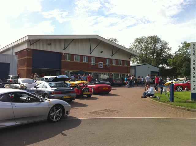 Time for some tyre kicking? - Page 1 - Northamptonshire - PistonHeads - The image showcases a parking lot in front of a large building with multiple garage doors underneath. The lot is filled with various cars, ranging from sports cars to sedans, all parked in an organized manner. Several people are scattered throughout the scene, some standing while others are sitting on the pavement, adding a sense of activity to the area. The setting, with its clear sky and open space, suggests a day enjoying outdoor activities.