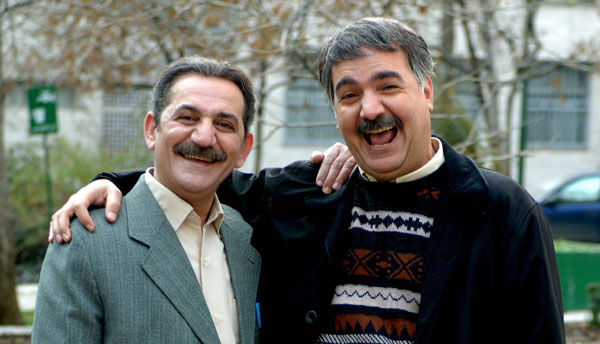 The image portrays two men in what appears to be a public outdoor setting. The man on the left is dressed formally, wearing a dark suit and tie. He has a surprised or amused look on his face. The man on the right is wearing a dark jacket over a striped sweater with a cream-colored shirt underneath. He appears joyful, laughing and holding the other man in an affectionate manner. Both men have smiles on their faces, and the man on the right has a visible 'rug' hair style. In the background, there are some trees and what might be a parked car or truck, partially obscured by the greenery.