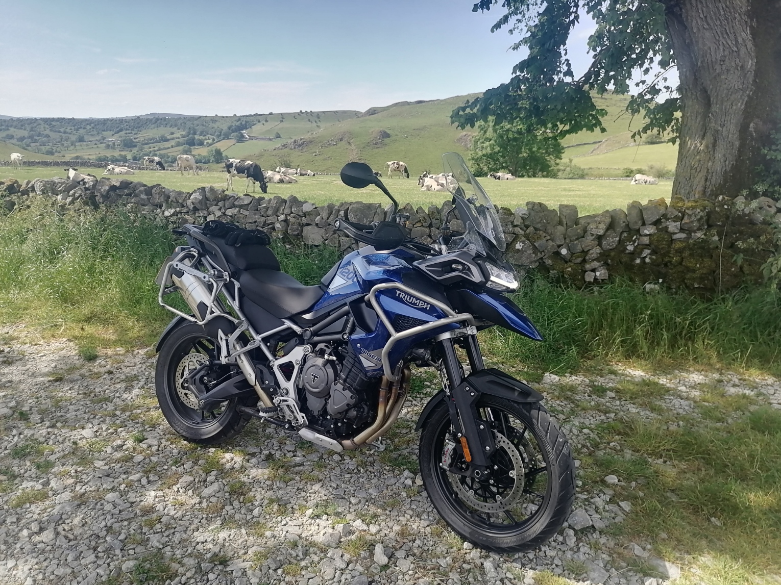 A picture a day...biker banter (Vol 6) - Page 133 - Biker Banter - PistonHeads UK - The image shows a blue and white motorcycle parked on a gravel surface next to a field. The bike has a prominent front wheel, a windshield in the center of the handlebars, and is equipped with what appears to be saddlebags on either side. In the background, there are rolling hills, and partially obscured by the motorcycle is a large boulder. The setting suggests a rural or countryside location with natural features like grass, stone, and trees visible.