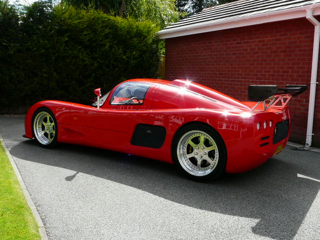 Who drives what? - Page 1 - East Anglia - PistonHeads - The image features a striking red sports car parked on a driveway next to a brick wall. The vehicle appears to be a convertible with a streamlined design and a long body. It's positioned at an angle, highlighting its profile and the broad stance of its wheels. The car's shiny exterior reflects the light, suggesting it may be in excellent condition or freshly polished. The setting suggests a private residence, with the driveway being the main area of interest.