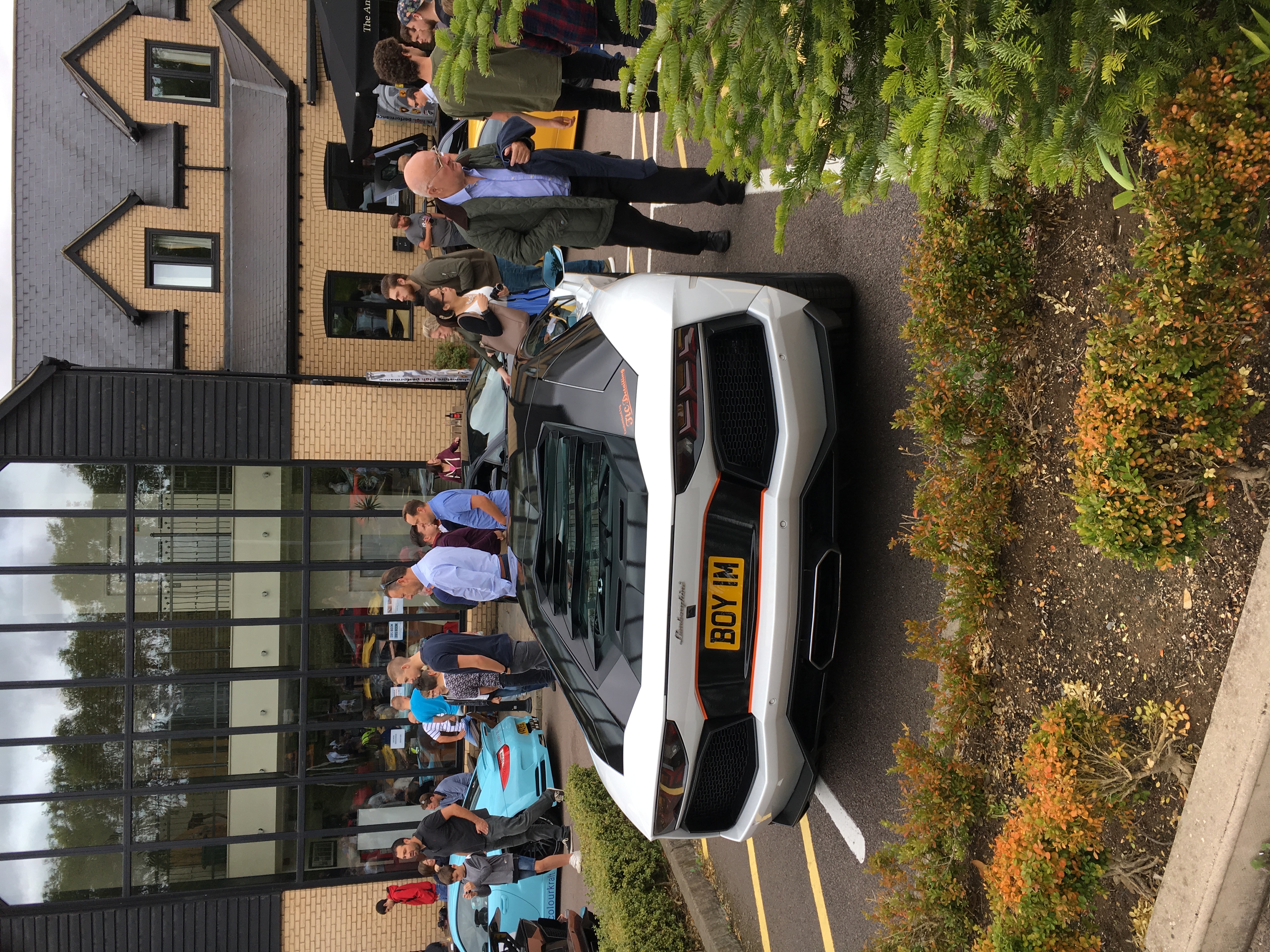 Sharnbrook today  - Page 1 - Events/Meetings/Travel - PistonHeads - The image depicts a scene where a white luxury car is parked in a street lined with businesses. Surrounding this car is a crowd of people, some of whom appear to be inspecting the vehicle. A black umbrella is open, providing shelter to a person standing near the car. The setting suggests this event might be part of a public gathering or showcase in a city.