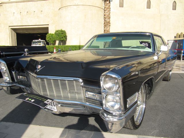 Classic Pistonheads Emaar - This image portrays a classic, black Cadillac car parked in front of a large, beige building. The car is positioned at a slight angle to the camera, allowing a view of its front and side. The vehicle is shiny and appears to be in good condition, suggesting it might be a restored vintage model. The setting seems to be a clear day as the car is casting a shadow on the ground, reinforcing the light source from above. The contrast between the black Cadillac and the beige building gives an aesthetic balance to the scene.