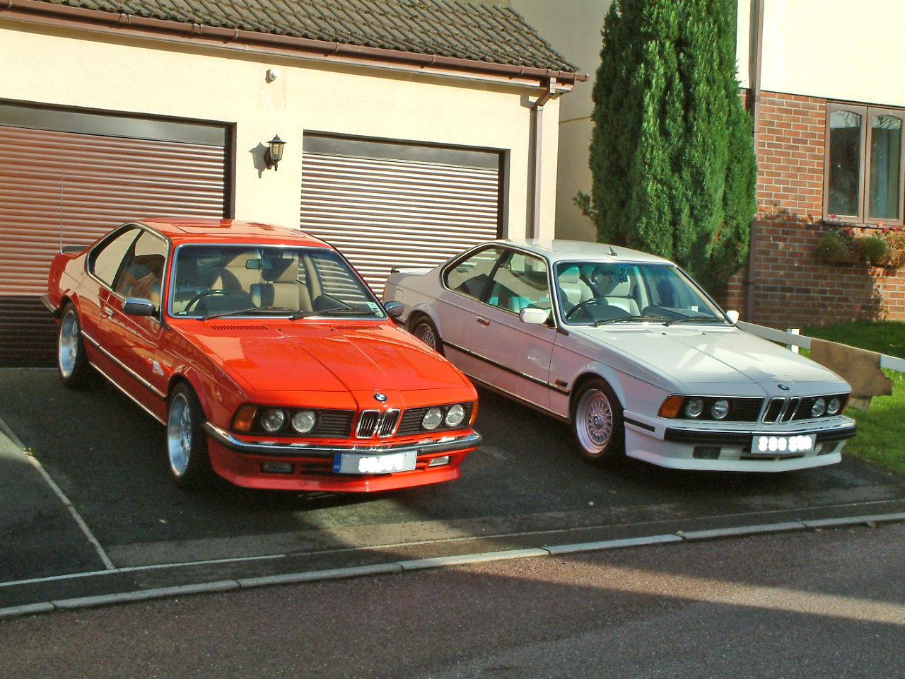 Six appeal - Show us your six! - Page 3 - BMW General - PistonHeads - The image showcases a formal front yard where two luxury cars, an eye-catching red R8 Audi and a white BMW 750 Li, are parked side by side in front of a garage. The garage door features a series of glass panels and a large window on its right side. A well-manicured lawn, a red brick wall, and a smattering of greenery along the right edge of the image enhance the suburban setting.