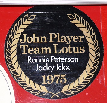 Show Us Your Stickers! - Page 1 - Formula 1 - PistonHeads - The image features a circular plaque design that is clearly antiquated, having elements like a clock at the bottom and text at various points within the circle. The prominent text at the top reads "JOHN PLAYER TEAM LOTUS" in capital letters. Below that, the names "RONNIE PETERSON JACKY ICKX" are displayed, with "RONNIE PETERSON" in a larger font size than "JACKY ICKX." 

At the bottom of the plaque, there is a digit "1975" which may denote a notable year, and it's situated in the lower part of the clock. The entire plaque is bordered by a decorative border that includes laurel leaves or a similar design.