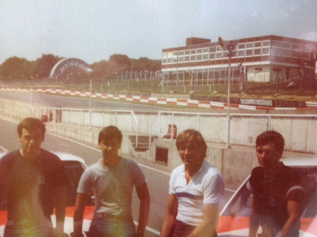 James Hunt/Marlboro Challenge /1983 XR3i  JNO 2Y - Page 1 - Classic Cars and Yesterday's Heroes - PistonHeads - In the image, there are four men standing together. They're positioned in front of a vehicle that spans the width of the frame, suggesting it might be a boat. Behind them, a fenced racetrack snakes through the landscape, with a building visible in the distance. The photo appears to be taken in daylight, under clear skies. The overall scene suggests an outdoor motorsport or recreation event.