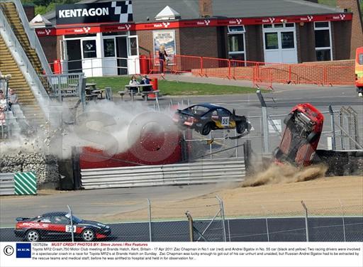 MR2 Championship this weekend... anyone else on here? - Page 4 - UK Club Motorsport - PistonHeads - The image captures a dramatic scene from a racing accident on a track near a large red structure, possibly a reception or administration building, identified as KENTGON. Two racing cars are prominently featured, one turned upside down and the other veered off course, suggesting a high-speed crash. The track is lined with a green and white fence, which is now in disarray due to the aftermath of the crash. The surrounding area is filled with debris from the wreckage, and spectators can be seen scattered around the track. A red building is in the background, presumably overlooking the speedway or restaurant near the track.