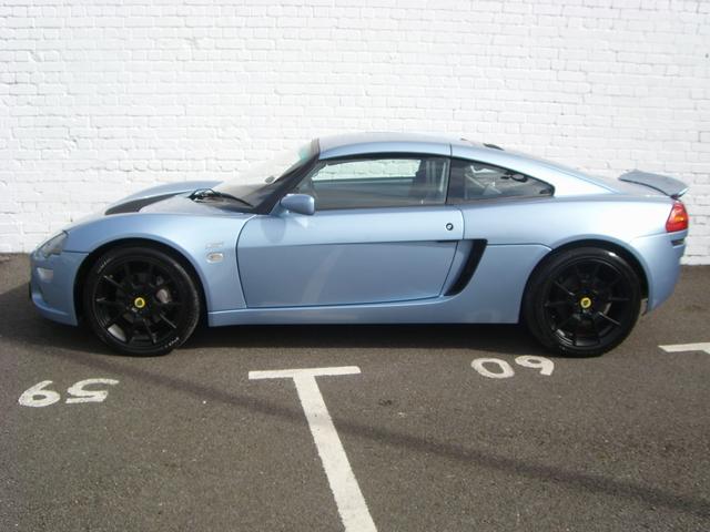 Europas - why such dowdy colours?! :-) - Page 1 - Elise/Exige/Europa/340R - PistonHeads - The image shows a silver sports car parked in a parking lot. The car is facing the camera and appears to be a two-door model, with the power window controls situated in the doors. It is parked at a parking space marked by the number "65" and the number "09" is visible at the front of the car. The background features a white wall behind the car, suggesting that the photo was taken in a controlled parking area or a parking garage.