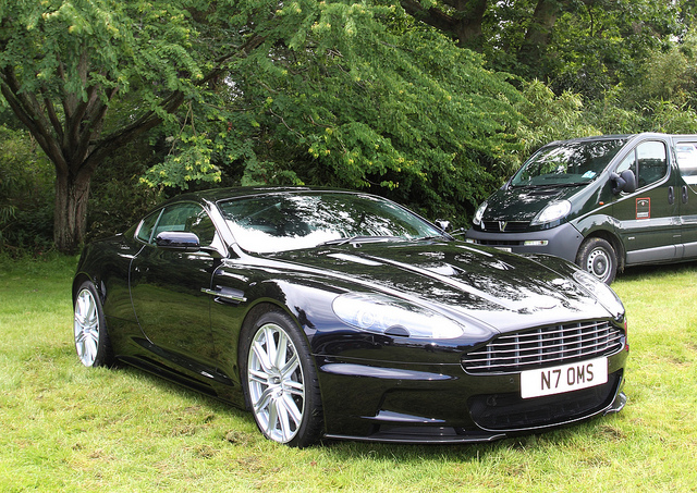 HEVER 2013  - Page 10 - Aston Martin - PistonHeads - This image shows a dark-colored luxury sports car parked on a grassy field in a park-like setting. The sports car is positioned in the foreground, taking up a large portion of the frame, with the hood prominently visible. In the background, slightly blurred to emphasize the car, is a silver and black SUV. The surrounding area is lush with trees, indicating a serene outdoor environment. The license plate of the sports car is clearly visible, among some other car details such as the headlights and one of the doors.