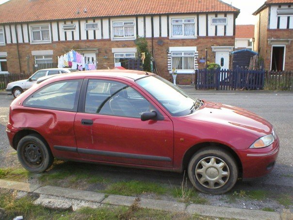 My Car - Page 1 - Readers' Cars - PistonHeads - This image shows a red car parked on a street alongside a row of terraced houses. The car features a sunroof, four doors, and a standard wheel encapsulating a hubcap without a central centerpiece. In the background, there's a green lawn and a fence, reflecting the typical urban suburban setting. The car appears to be in good condition apart from the visible scratches on the side. The overall lighting suggests it could be an overcast day. The street has a laid back atmosphere with minimal traffic on the road visible in the background.