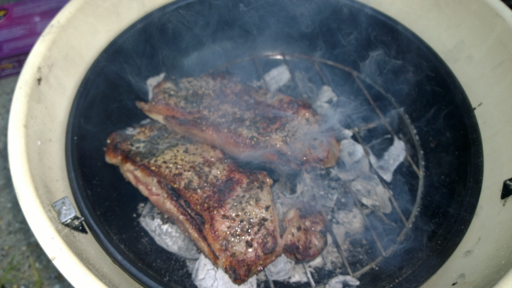 THE  STEAK THREAD, served a la Man. - Page 10 - Food, Drink & Restaurants - PistonHeads - In the center of the image, there's a grill smoker where several pieces of food, possibly cuts of meat, are being cooked under a moderate amount of smoke. The meat has a well-seasoned appearance and appears to be partially cooked with slightly charred edges. The grill is inside a larger metal container, which could be the body of the smoker or a protective cover. To the right of the image, there's a hint of a purple and white object that could be a piece of fabric or a package, but it's not clear due to the angle and cropping of the image.