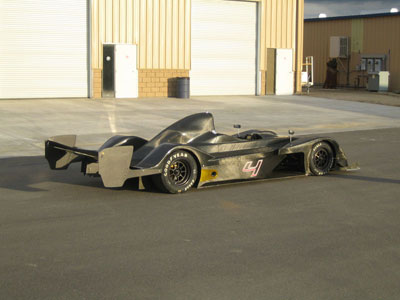 Pistonheads - The image shows a sleek, streamlined racing car sitting idle on a concrete pavement. The car is colored in shades of black and grey with subtle red accents, adding to its sporty aesthetic. Its design features large, open wheels and a low-slung body, indicative of its purpose-built functionality. The car is parked outside a garage or warehouse with large roll-up doors, suggesting a setting typically used for vehicle storage or manufacturing. The background is minimally decorated, with scattered clouds hinting at the possibility of an overcast or cloudy day.