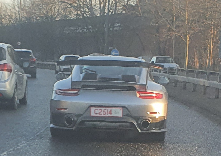 A car is parked on the side of the road - The image shows a gray sports car on the road. It has a visible license plate and an open sunroof. The vehicle's rear wing suggests it is designed for high-speed driving or racing, and its sleek design implies a focus on aerodynamics and performance. The background includes a highway with multiple lanes, a guardrail, and other vehicles in motion, indicating that this photo was taken during a drive. There are no visible texts on the car itself that provide additional context.