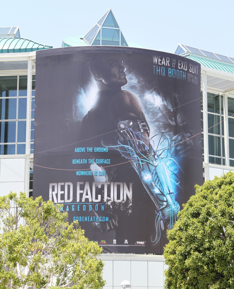 The image shows a promotional banner of a movie against a backdrop of a modern outdoor building, which seems to be made of glass. The banner prominently features the title "RED FACTION" and attributes the roles of the characters: "Godfather," "Queen of Ice Cream," and "Samurai." The artwork on the banner depicts an action-packed scene with a superhero dressed in a black suit with light effects, suggesting an exciting and high-energy movie. The sky above appears clear, suggesting good viewing conditions for the banner.