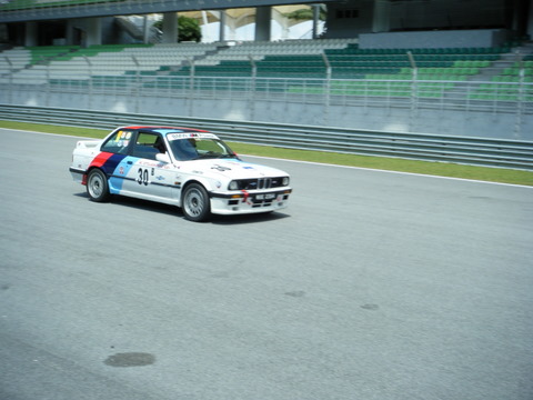 Afos Sepang Pistonheads - In the image, a vintage car is the star of the show. It's sporting a vibrant livery of blue, red, and white, painted in abstract fashion, giving it a dynamic and energetic look. The car is part of a motor race, as evidenced by the race track and concrete barriers in the background. A large crowd fills the bleachers, keenly watching the adrenaline-fueled action on the track. The car's number, 30, is prominently displayed, indicating its participation in the race. The overall scene is filled with anticipation and excitement.